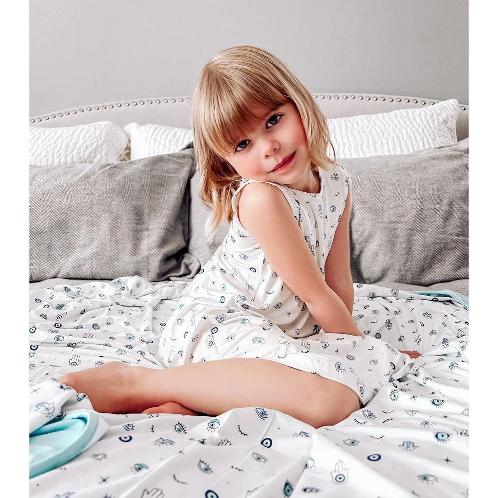 Bellabu Bear The Eye Bamboo Girls Sleeveless Dress, white with blue eye print, worn by a smiling toddler girl on a bed.