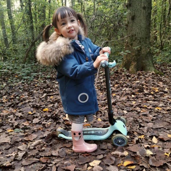 Boxbo Wistiti Fleece Lined Rainboots Glitter Pink; toddler girl in a blue coat stands on fallen leaves, riding a pastel scooter.
