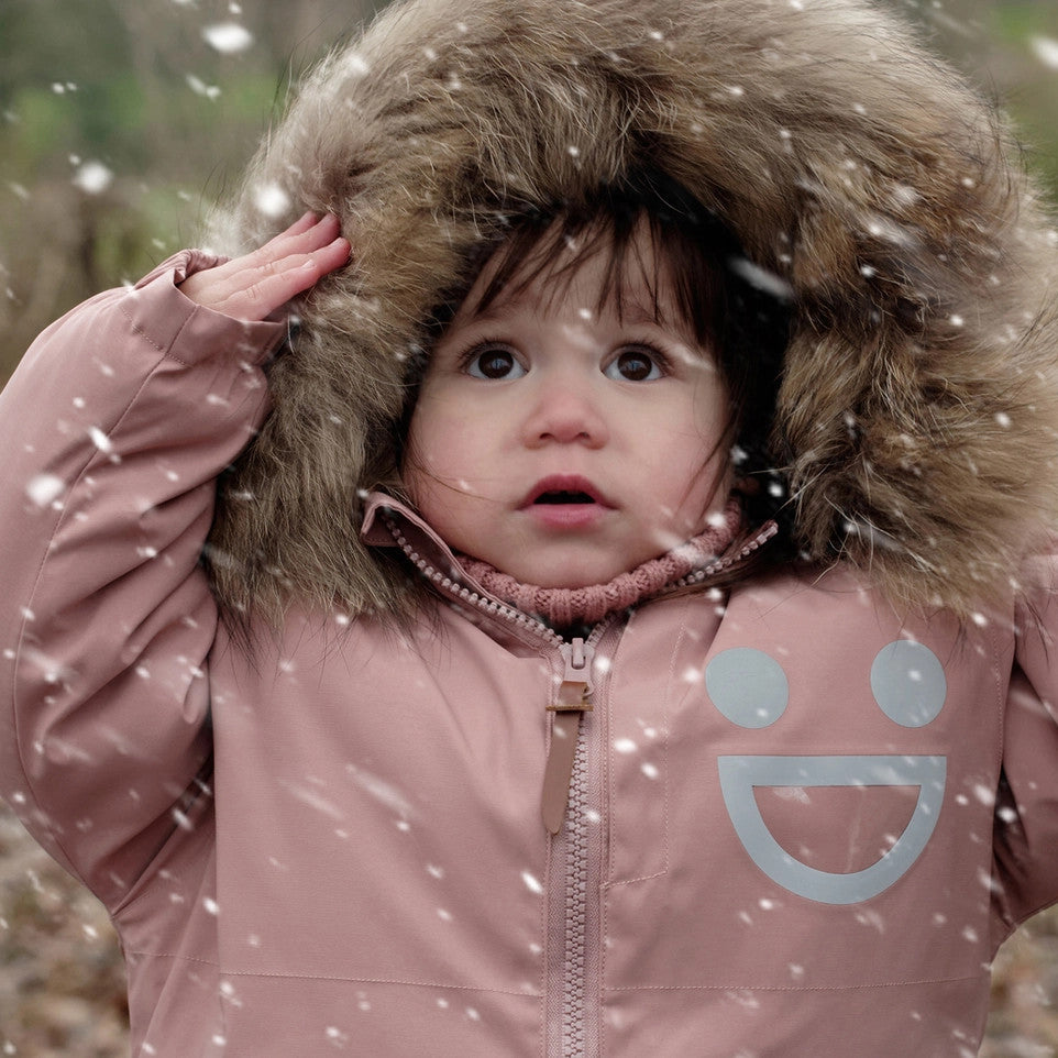 Boxbo Wistiti fleece-lined waterproof fur jacket for toddlers in dusty rose pink, child adjusting fluffy faux-fur hood in falling snow.
