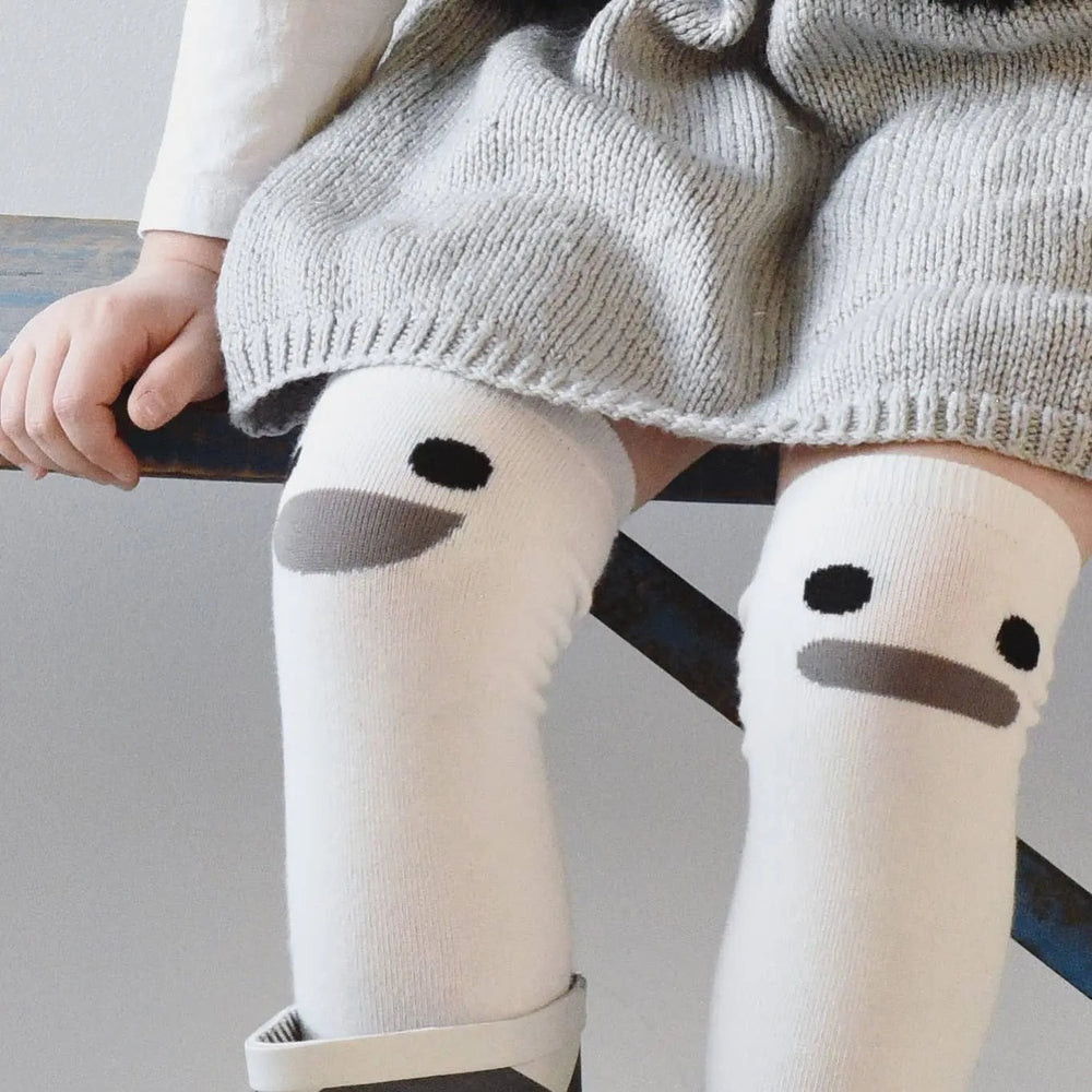 Boxbo Wistiti High Sock Blanc, white knee-high socks with black eye and gray beak on a baby sitting on a bench