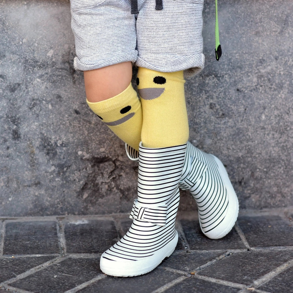 Boxbo Wistiti High Sock Yellow, yellow smiley-face socks shown above striped rain boots as a toddler stands.