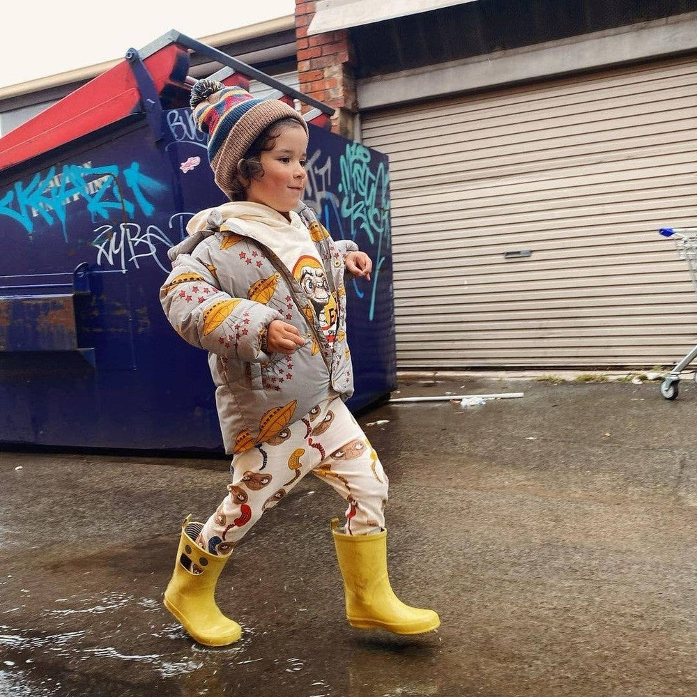 Boxbo Wistiti Rainboots Moutarde: mustard yellow kids' rain boots worn by a child walking on wet pavement in a graffiti-filled urban scene