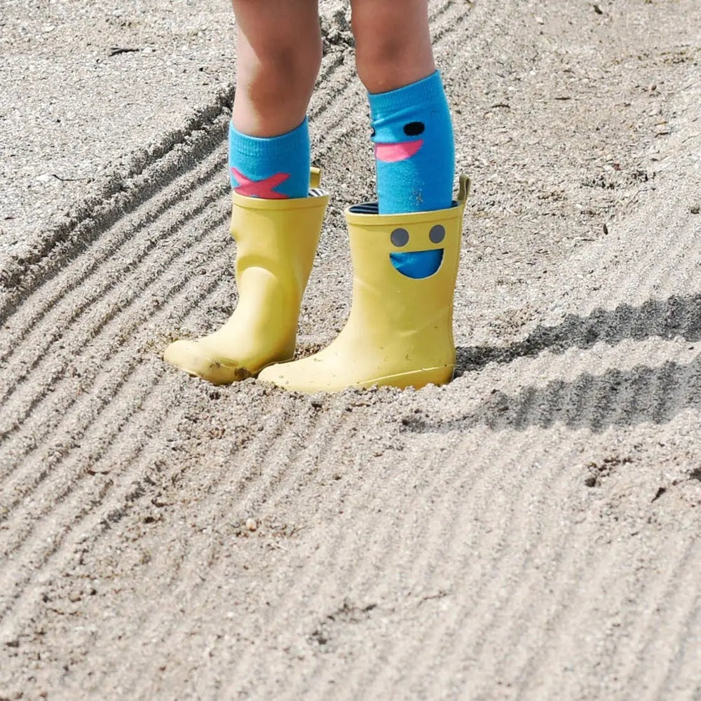 Boxbo Wistiti Rainboots Moutarde; yellow rubber boots with a smiling face, worn by a child standing in sand with blue socks