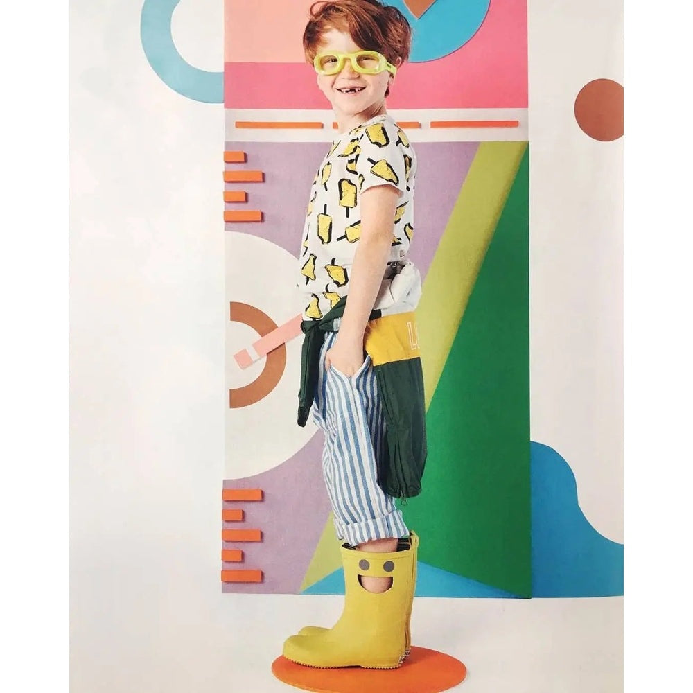 Boxbo Wistiti Rainboots Moutarde; mustard yellow boots with a smiley face worn by a smiling child.