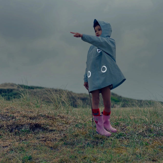 Boxbo Wistiti Windbreaker Rain Jacket Blue worn by a child outdoors with hood up, pointing in a windy field.