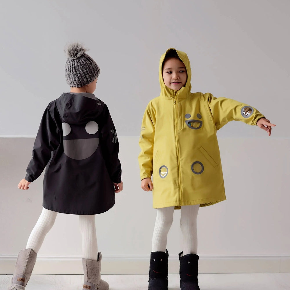 Boxbo Wistiti Windbreaker Rain Jacket Yellow worn by a smiling child with hood and outstretched arm; another kid in black coat beside.