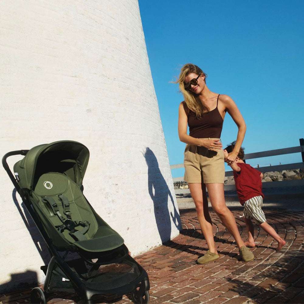Bugaboo Butterfly 2 stroller, green with harness, beside a white wall as mom and child walk.