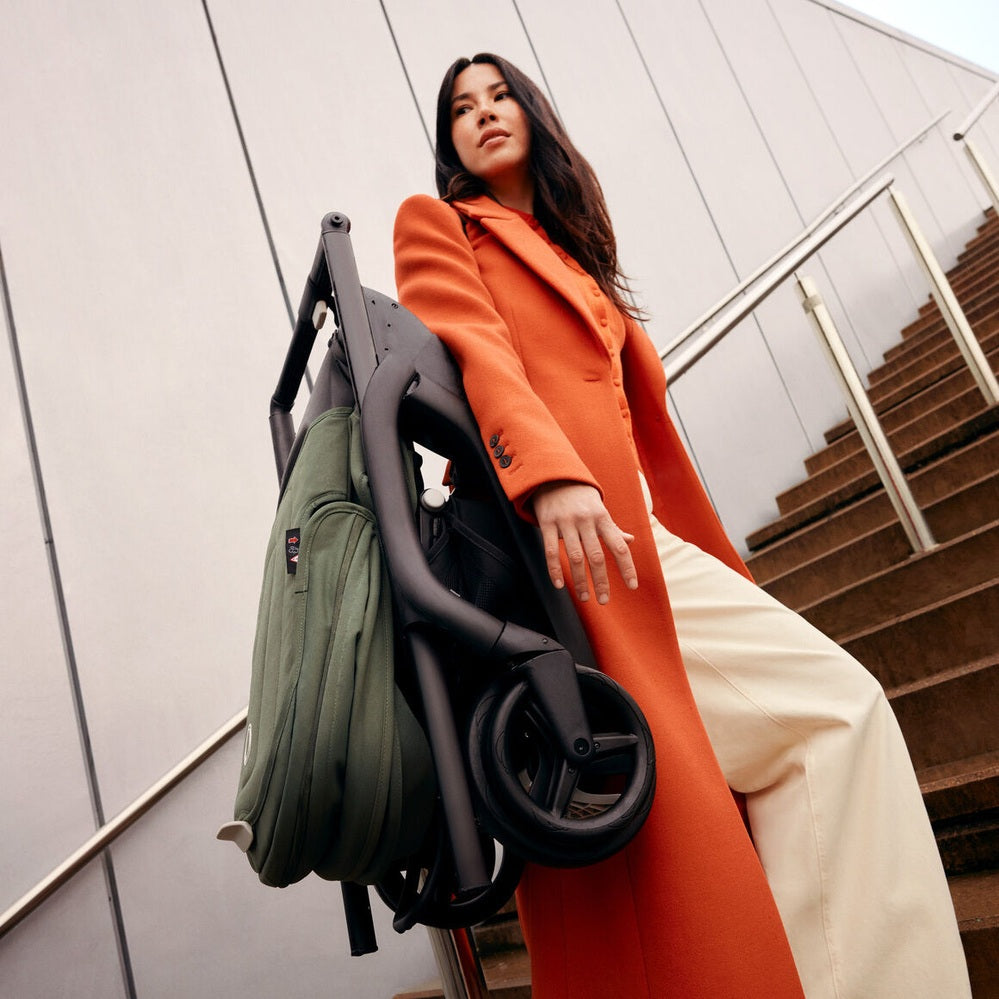 Bugaboo Dragonfly Stroller Complete folded in black frame with green bag, carried by a woman in an orange coat.