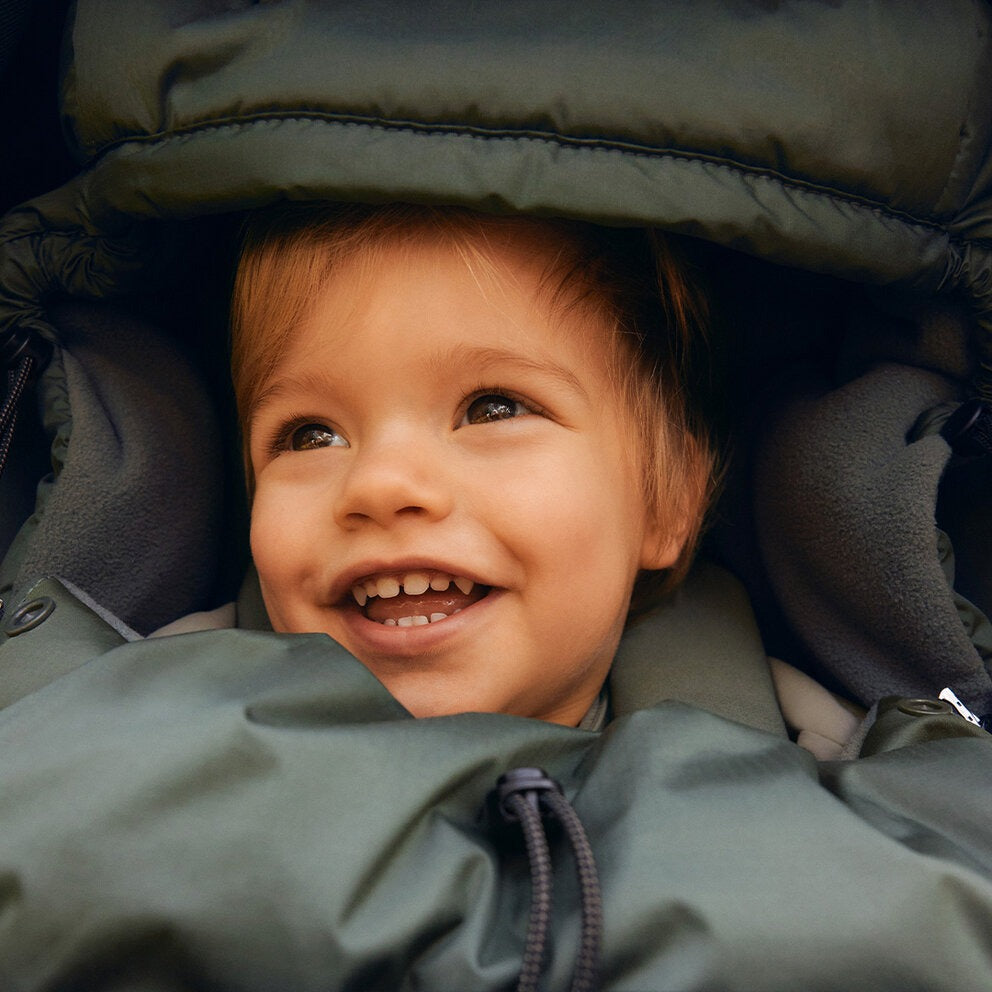 Bugaboo THERMOLITE Performance Footmuff in green, keeping a smiling toddler warm in a stroller.