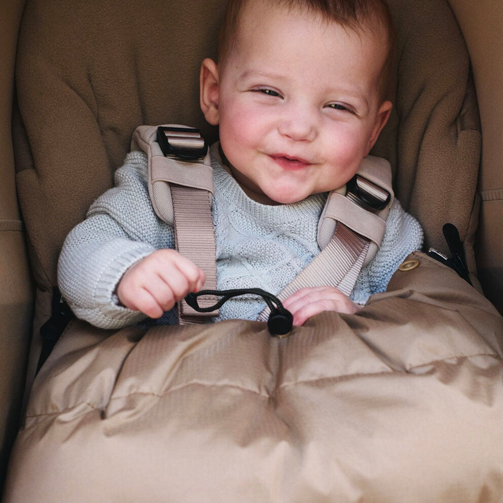 Bugaboo THERMOLITE Performance Footmuff in beige keeps a smiling baby warm, seated securely in a stroller.