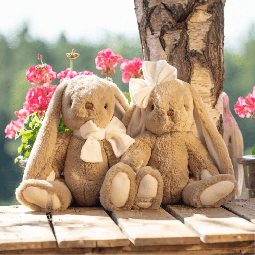 Bukowski Bears Bunny Patrick stuffed animals pair, beige with cream bows, sitting together on a wooden deck near pink flowers.