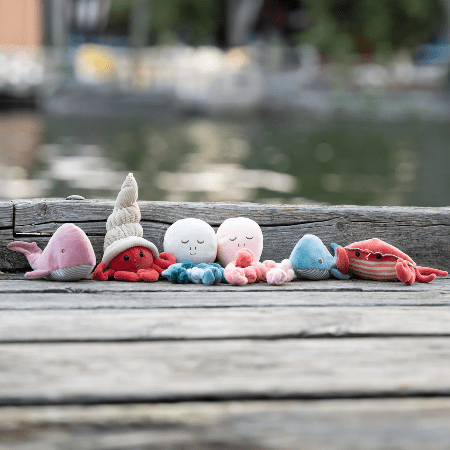 Bukowski Bears Hermit The Crab stuffed animal among colorful sea plushies on a weathered dock beside calm water.