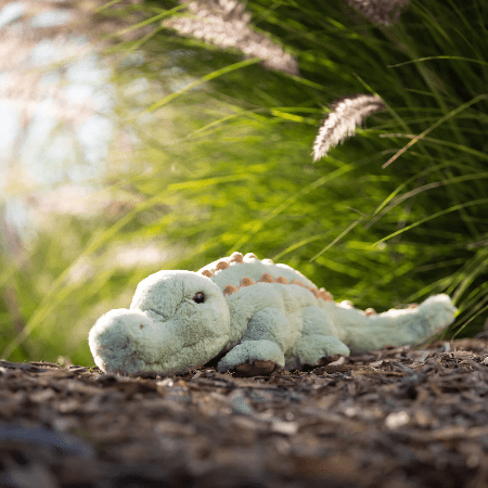 Bukowski Bears Maximus stuffed animal plush, mint green with tan spikes, lying peacefully on dirt amid sunlit garden grass.