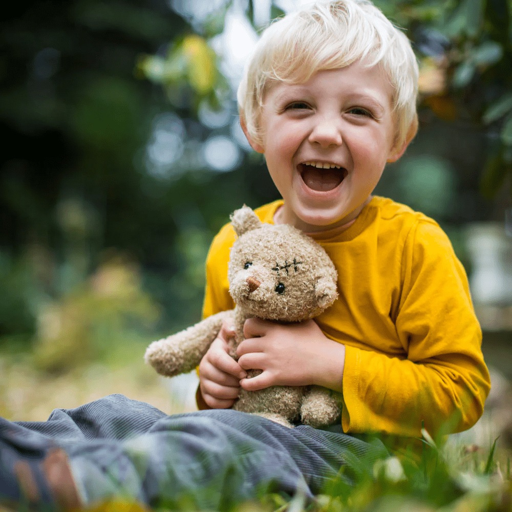 Bukowski Bears Nobody is Perfect stuffed animal; a smiling child in a yellow shirt hugs a beige plush bear outdoors.