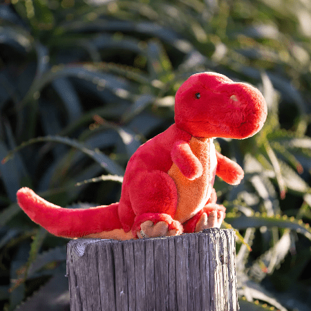 Bukowski Bears Rexie Dino stuffed animal, bright red plush with orange belly, perched on a weathered wooden post outdoors.