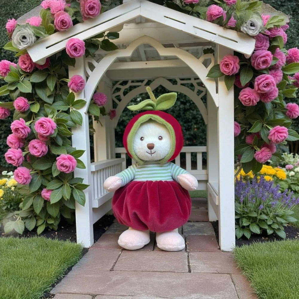Bukowski Bears Ziggy Cherry stuffed animal in a green hood and red velvet dress posed inside a white garden arch amid pink roses.
