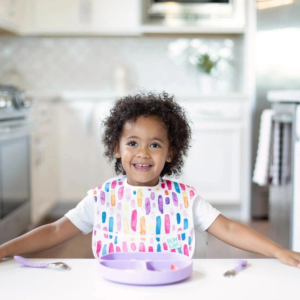 BUMKINS Silicone Grip Dish in purple sits firmly on a white table as a smiling toddler enjoys mealtime