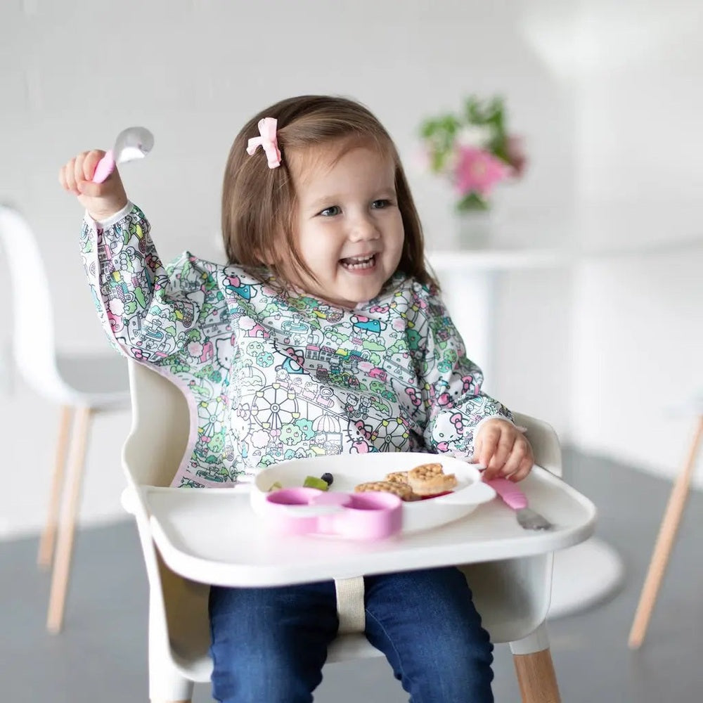 BUMKINS Sleeved Bib in colorful cartoon print; toddler enjoys mealtime with spoon, sleeves catching spills.