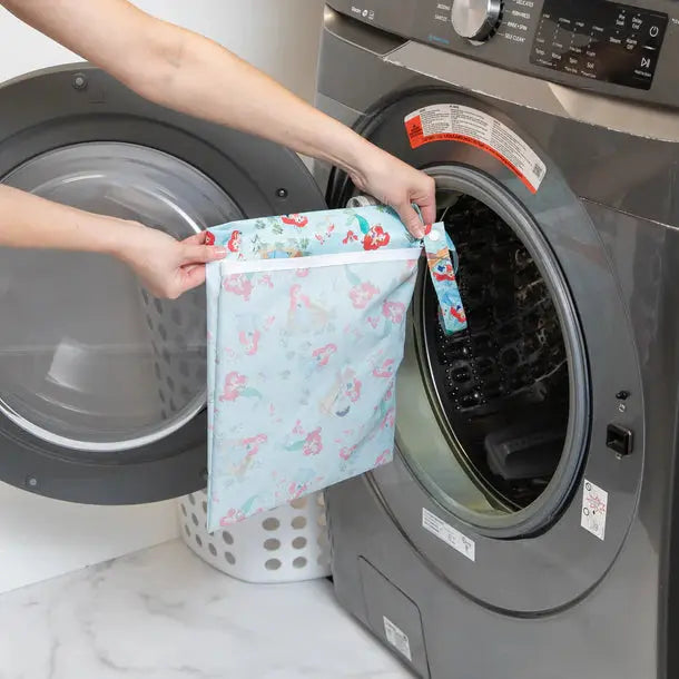 BUMKINS Wet Bag in light blue mermaid print, reusable diaper wet/dry bag shown being placed into a front-load washer.