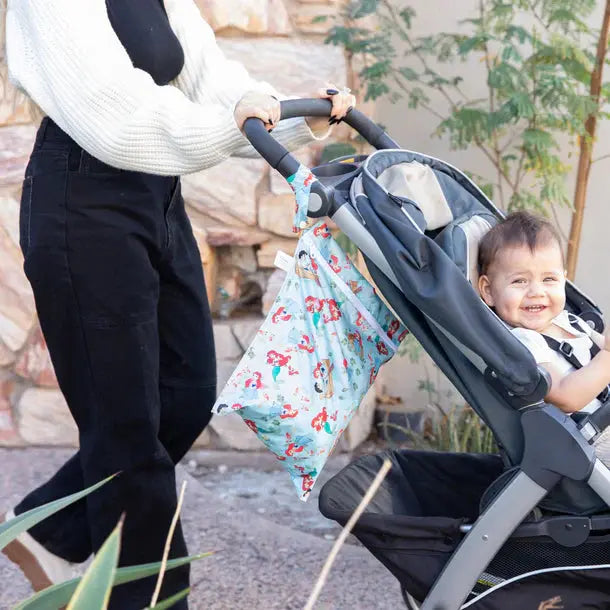 Bumkins Wet Bag in light blue with colorful print attached to a stroller handle as a parent pushes a smiling baby.