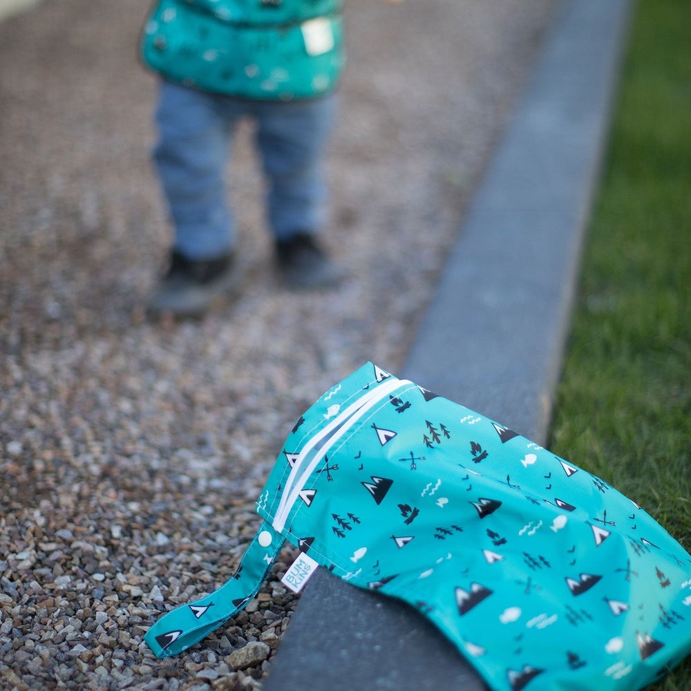 Bumkins wet bag turquoise with woodland tree print on gravel beside a child