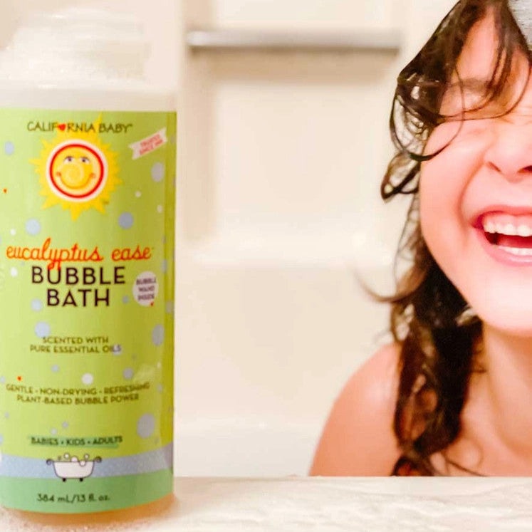 California Baby Bubble Bath Eucalyptus Ease 13 oz bottle next to a smiling child in the bath, green label visible