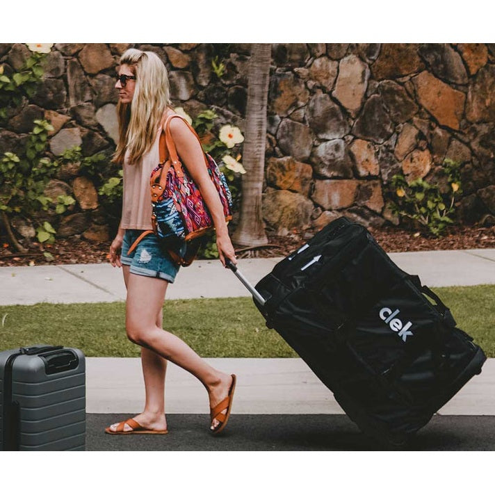 Clek Weelee baby travel bag on wheels; large black rolling bag with CLEK logo pulled by a woman in sunglasses