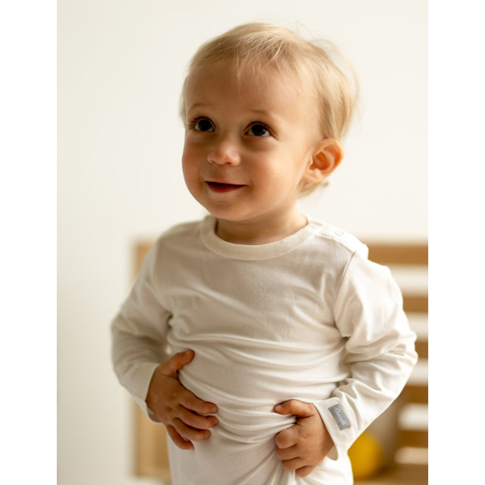 Coccoli Infant Cotton TENCEL™ Modal Long Sleeve Bodysuit Cream; smiling baby grips the cream outfit at the waist.