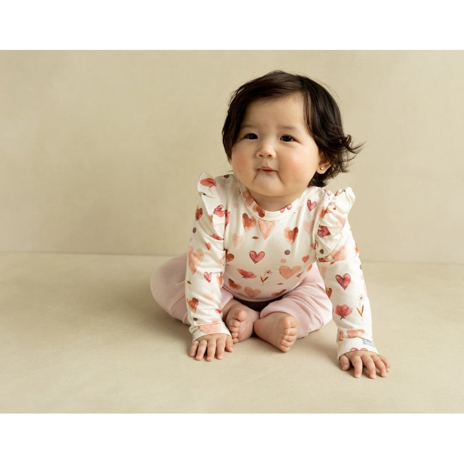Coccoli Infant Cotton TENCEL™ Modal Romper Hearts On Cream; baby sits on beige backdrop in a cream romper with pink hearts.