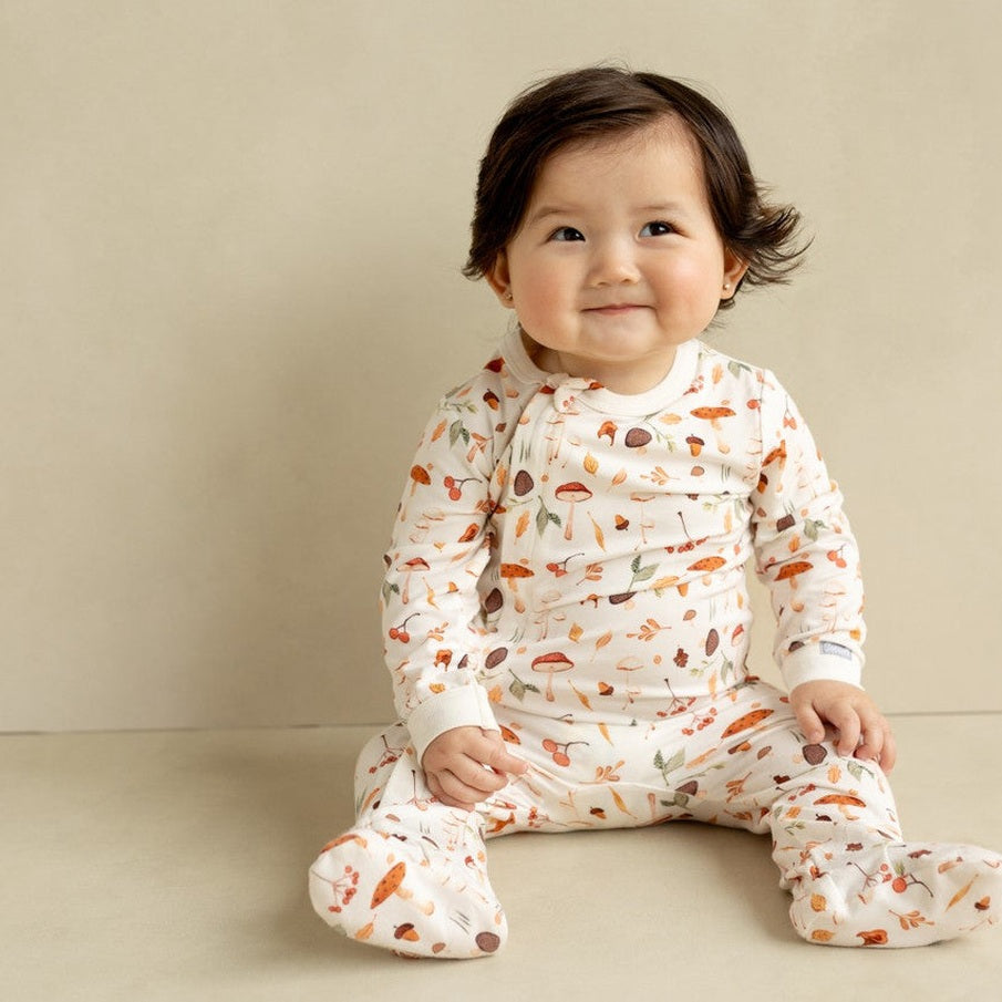 Coccoli Infant Cotton TENCEL™ Modal Zipper Footie Mushrooms On Cream; baby sits smiling in mushroom-print cream sleepsuit.