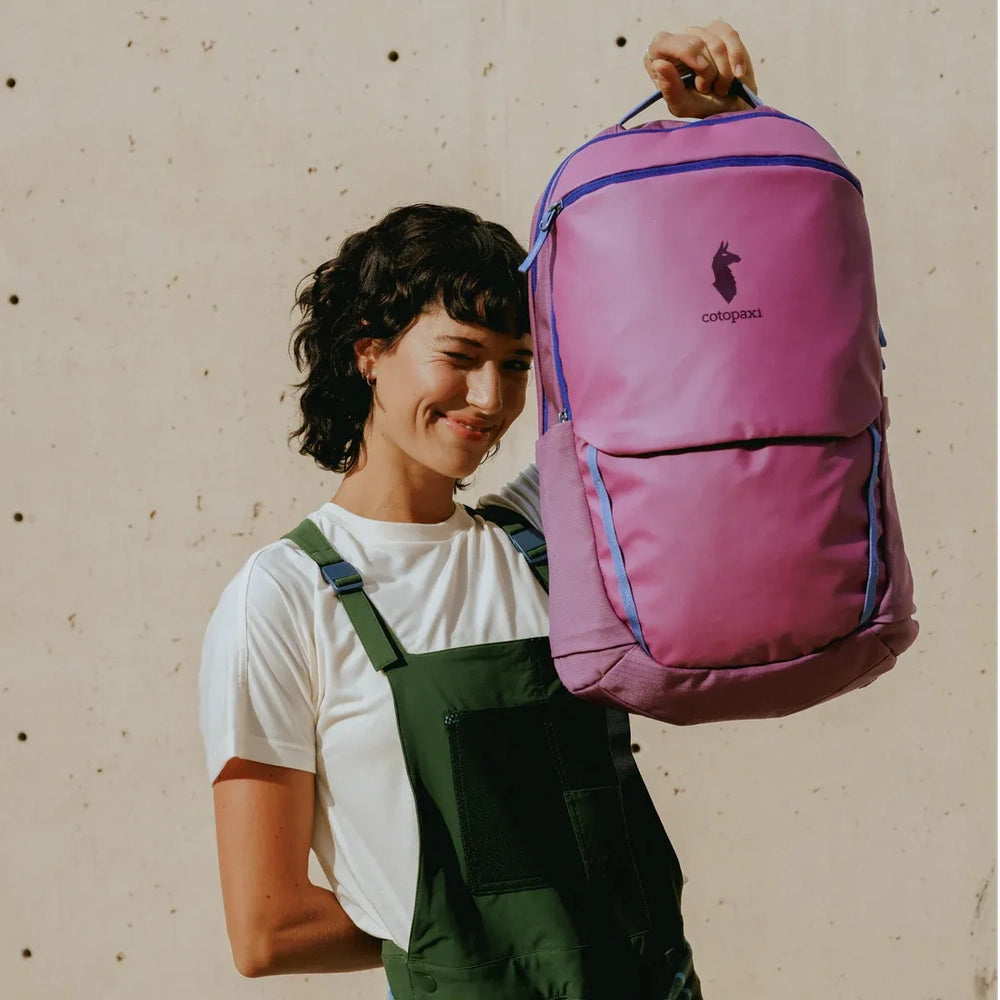 Cotopaxi Allpa 26L Daypack in pink-purple with blue zipper trim, held up by a person in white tee and green overalls.