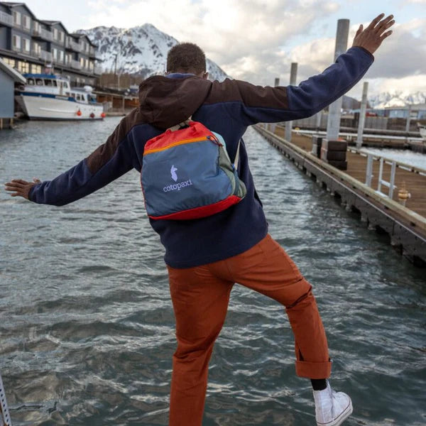 Cotopaxi Taal Convertible Tote in blue with orange top and red bottom, worn on back by person at marina.