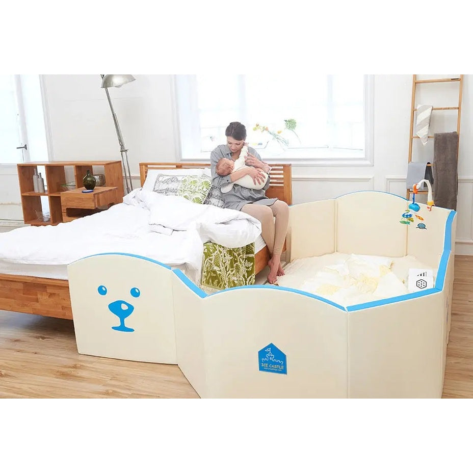 Creamhaus Grand Ice Castle Up BT baby play yard with blue trim and padded cream sides, mom cuddling baby beside it.