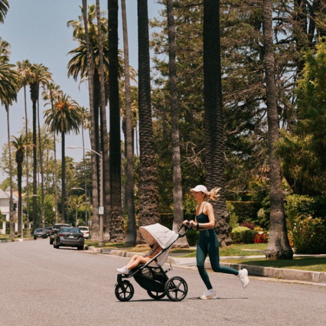 Cybex Avi Spin All Terrain Stroller in black frame with beige seat being pushed by a jogger along palm-lined street.