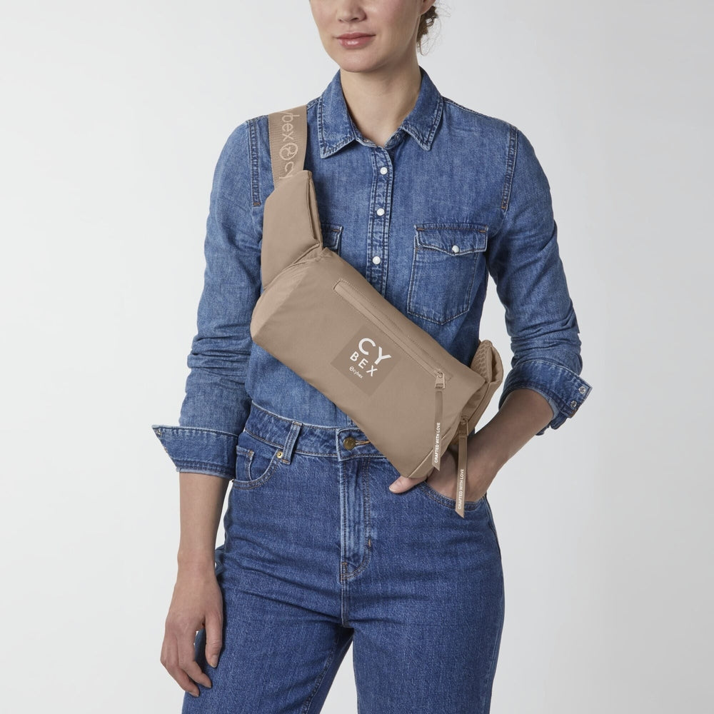 Beige Cybex Coya baby carrier worn crossbody with tan strap and front zip pocket.