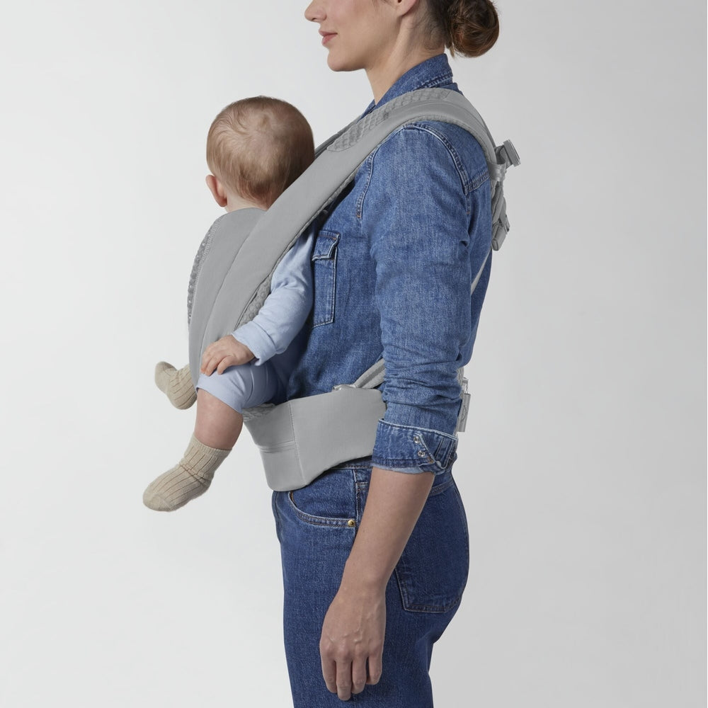 Soft gray fabric baby carrier worn by a denim-clad woman; infant in blue sits in front with padded straps and waistband.