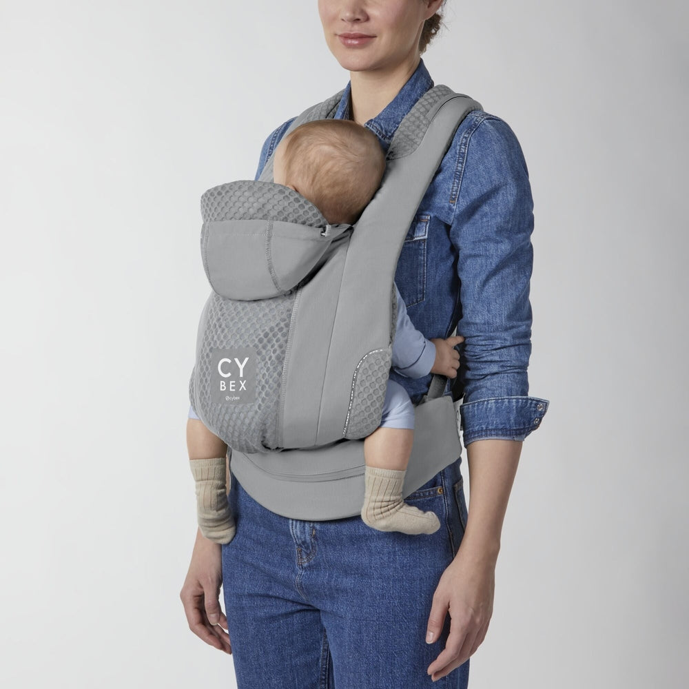 Gray Cybex Coya baby carrier with quilted mesh padding and hood, worn by a mom in a denim shirt.
