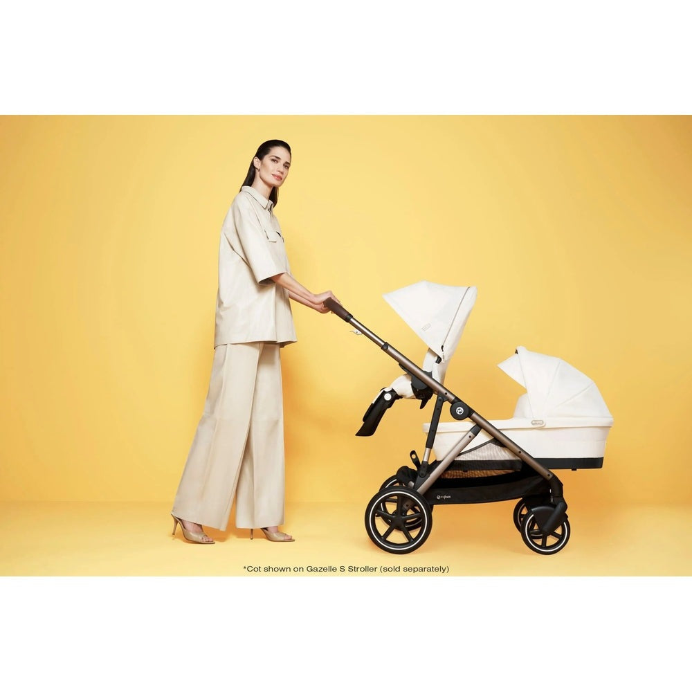 CYBEX Gazelle S V2 Cot stroller in beige with bassinet, being pushed by a woman on a yellow backdrop.