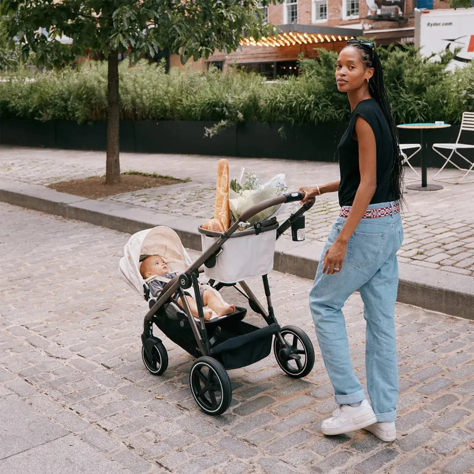 CYBEX Gazelle S V2 Stroller, black frame with gray fabric, mom pushes on cobblestone street with baby aboard