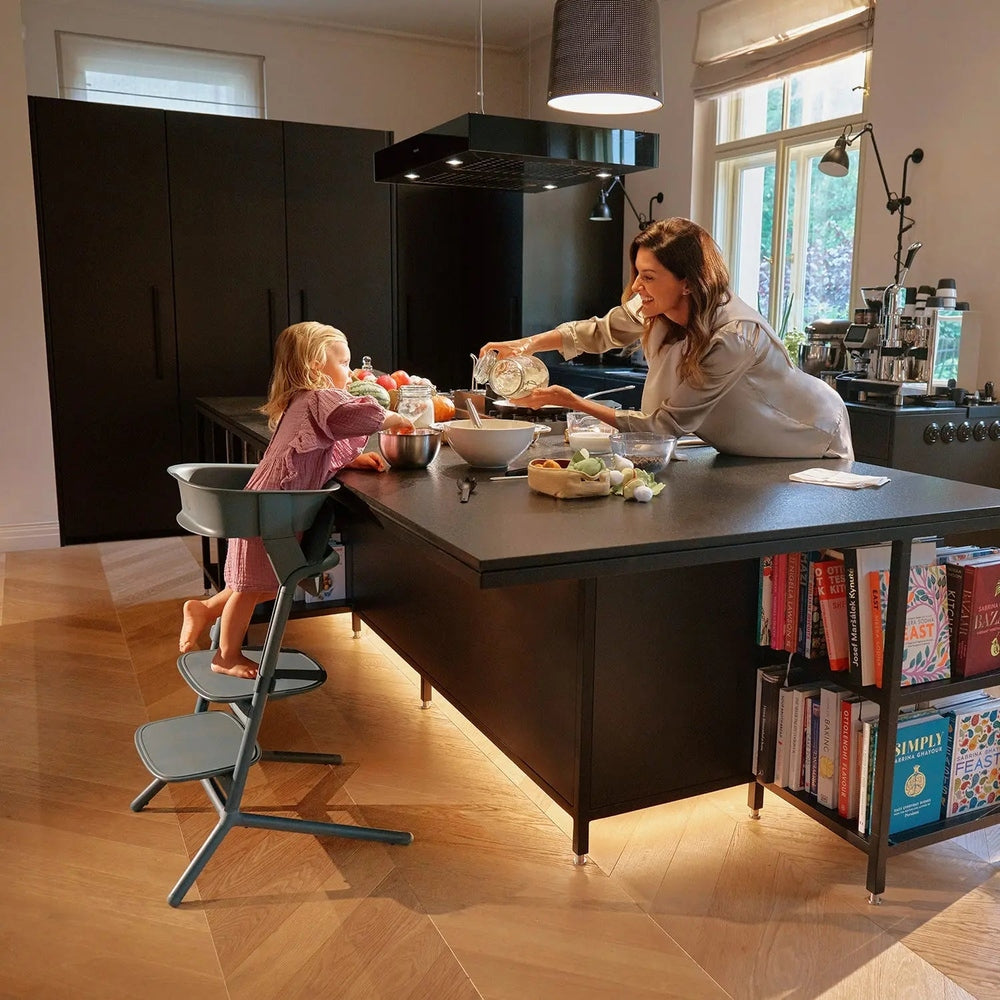 Cybex Lemo Training Tower in gray-blue at a kitchen island, child seated and reaching bowls while caregiver cooks.