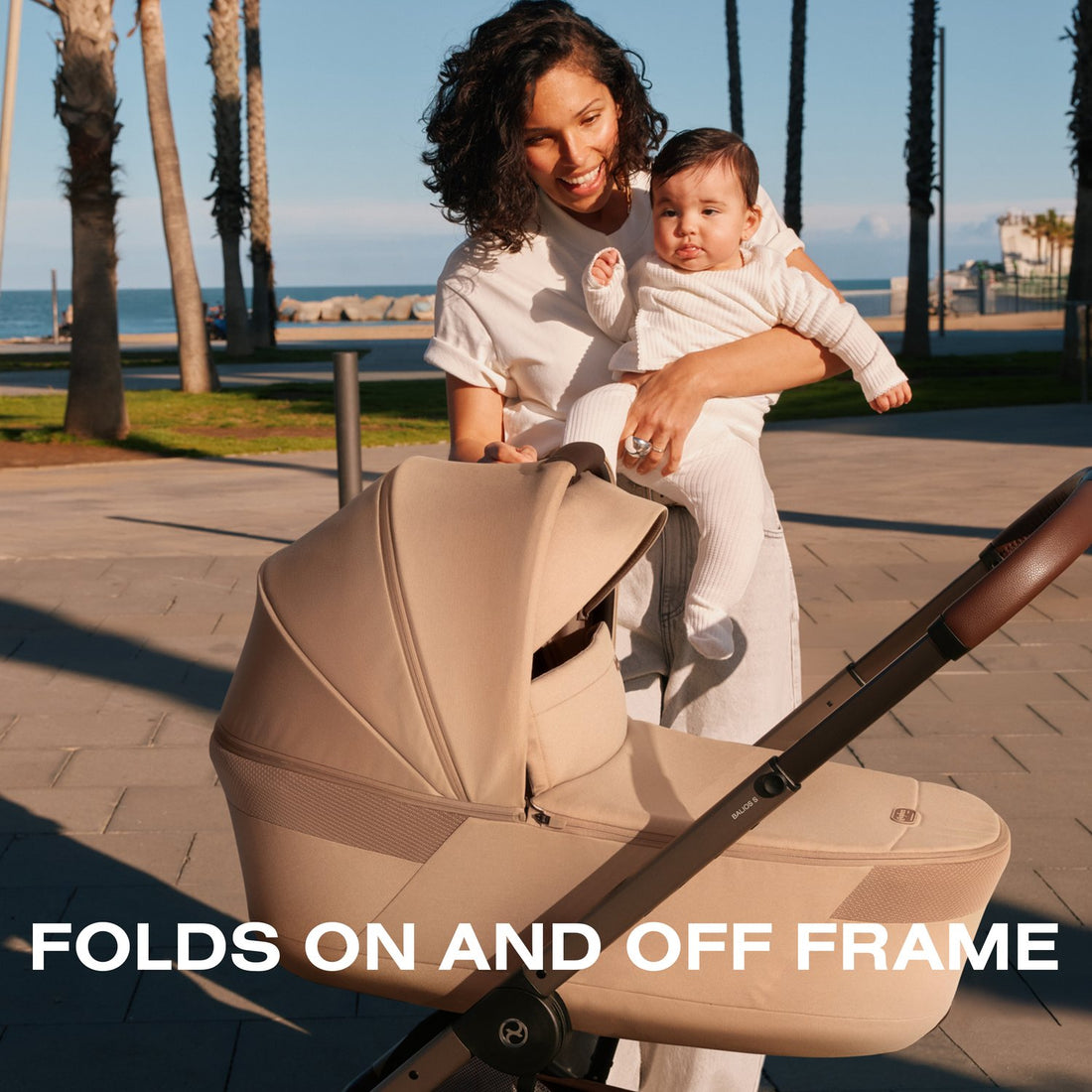 Almond Beige Cybex Balios Fold Cot on its stroller as a mother holds her baby on a palm-lined promenade