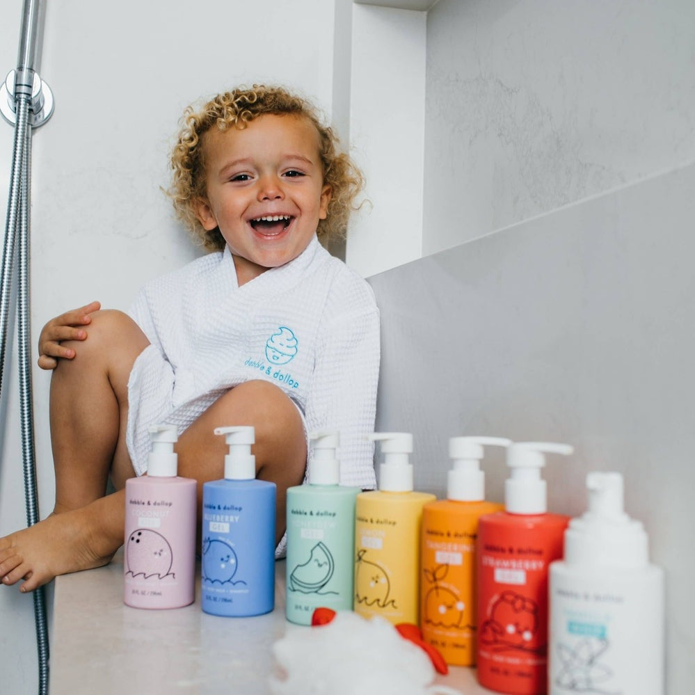 Dabble & Dollop All-Natural Shampoo, Body Wash & Bubbles Blueberry lineup as a joyful child sits on the tub edge in a bathrobe.