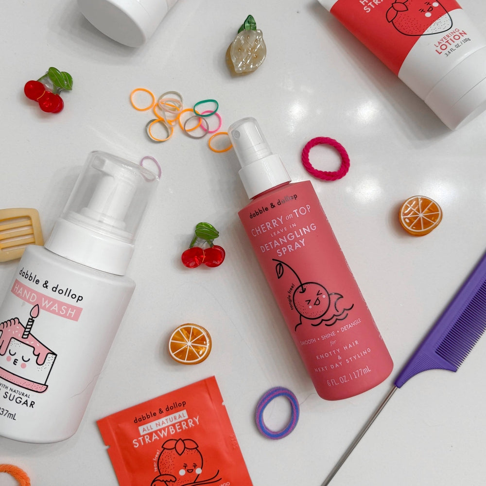 Dabble & Dollop Cherry on Top Detangling Spray in pink bottle with spray nozzle, beside a purple comb and hair ties.
