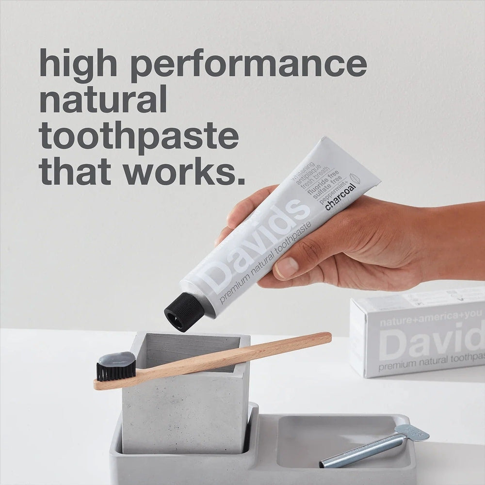 Davids toothpaste Fluoride Free Charcoal Peppermint being squeezed onto a bamboo toothbrush in a minimalist setup.