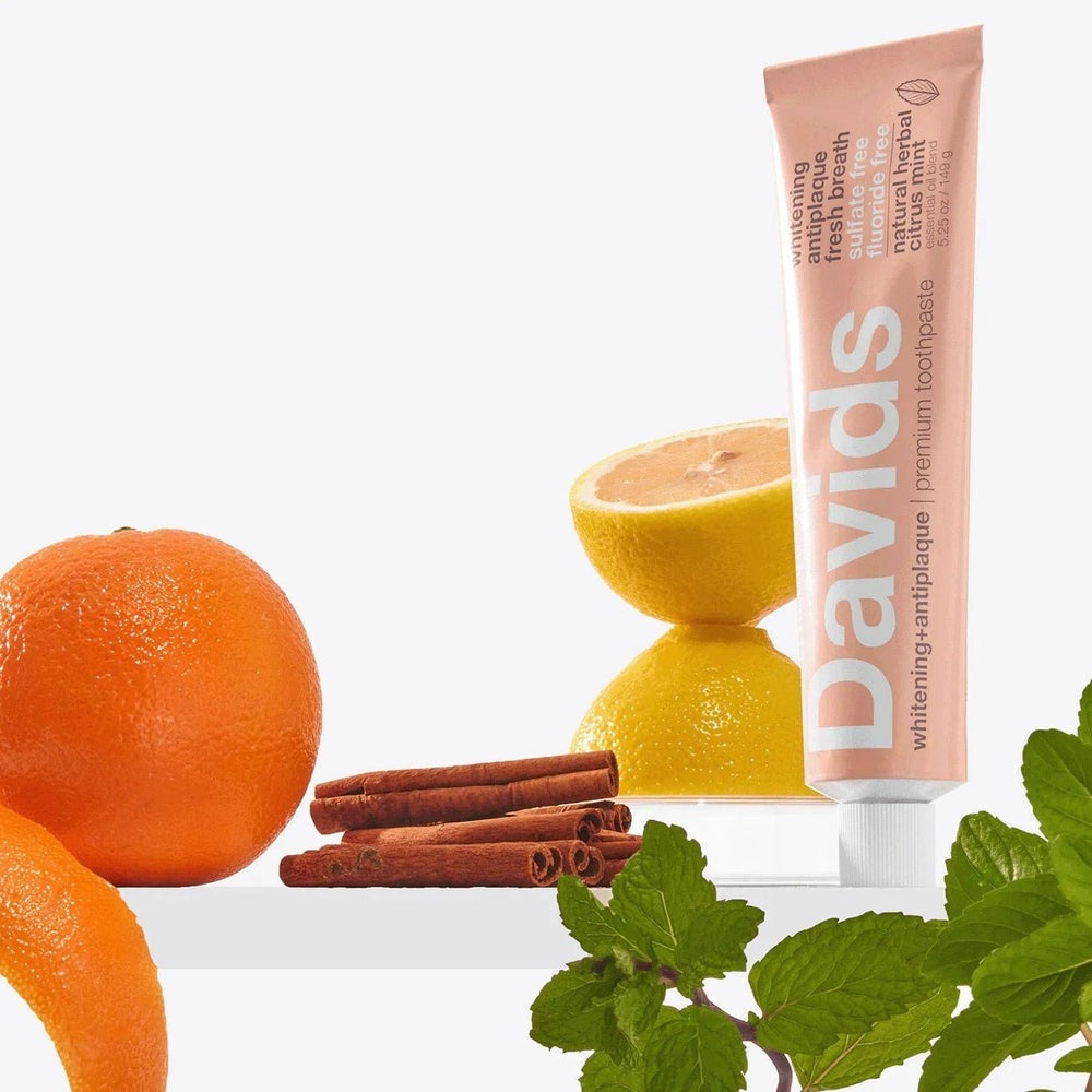 Davids Toothpaste Fluoride Free Herbal Citrus Peppermint tube stands upright beside oranges, lemon, cinnamon and mint.
