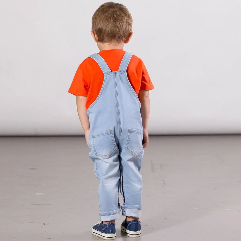 Deux Par Deux French Terry Denim Overalls Unisex in light blue; toddler backs to camera, orange tee visible, back pockets shown.