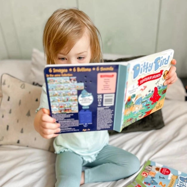 Ditty Bird Sound Book Dinosaur Sounds USB-C Rechargeable, a child sits on a bed reading and exploring sounds.
