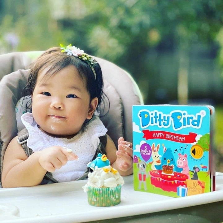 Ditty Bird Sound Book Happy Birthday USB-C Rechargeable beside a smiling baby in a high chair with a cupcake on the tray.