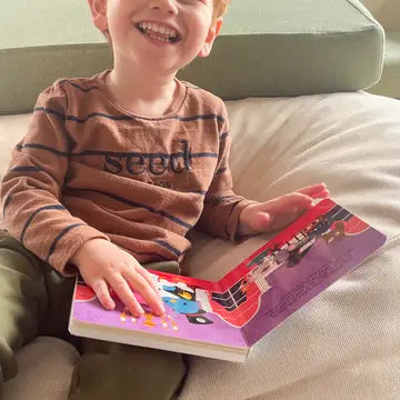 Ditty Bird Sound Book Music to Dance To USB-C Rechargeable; smiling child sits on a couch holding the colorful book.