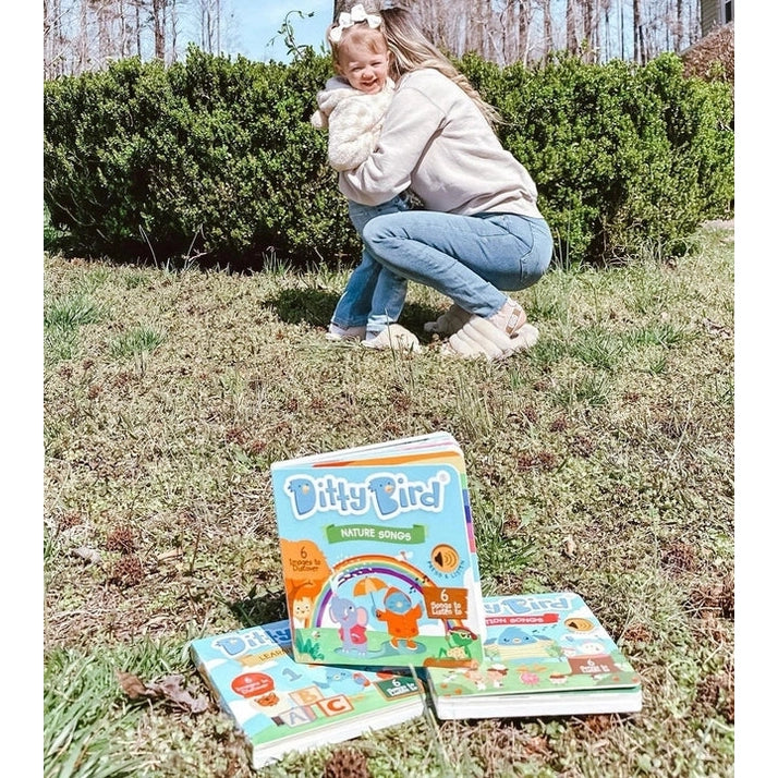 DITTY BIRD Sound Book Nature Songs USB-C Rechargeable enjoyed outdoors as a mother hugs her toddler beside colorful books.